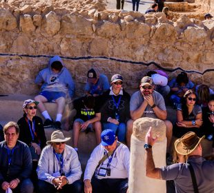 Group photo of tourists sitting under the shade of a wall.
