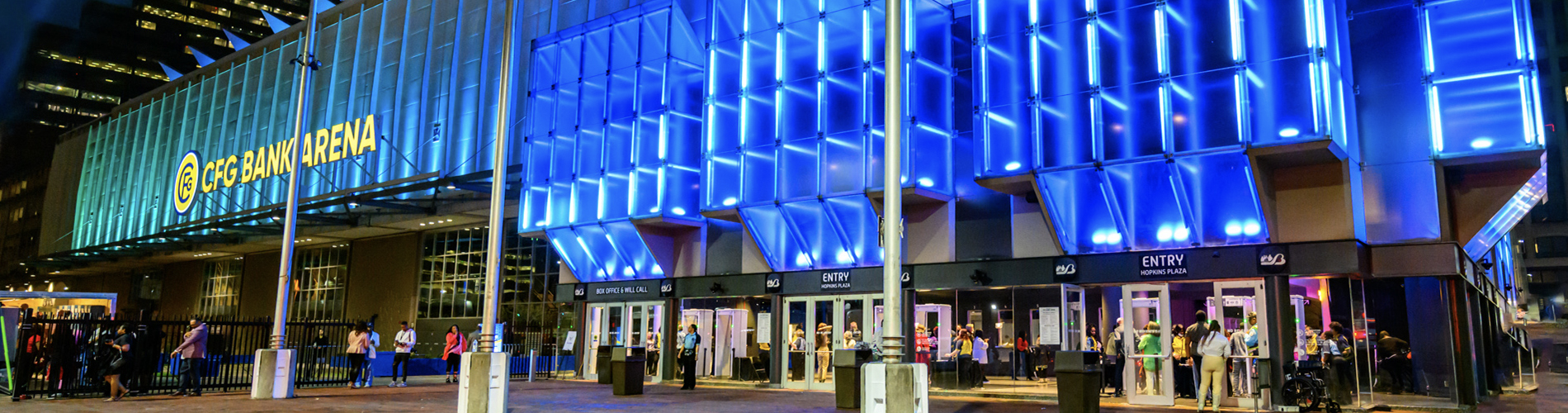 image of the CFG Bank Arena at night. The arena facade is lit with blue lights as people are coming in and out of the entrances.