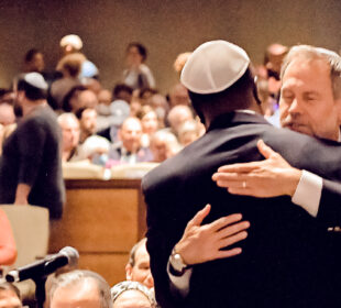 Two man are in the foreground hugging each other. One man's back is facing us, he is wearing a black suit and a kippah, the other man is facing the camera. There are a crowd of people behind them.