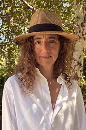 Photo of Beth Ricanati looking into camera. She is wearing a white shirt and a tan brimmed hat. There is a tree in the background.